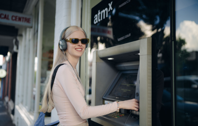 A blonde woman in a pink top, headphones and sunglasses smiles at the camera while using an ATM.