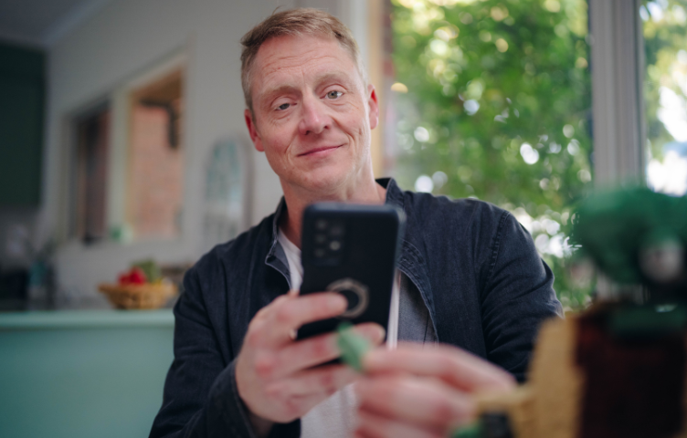 A blonde man in a blue shirt and white t-shirt sits at his kitchen table, looking at his phone screen.