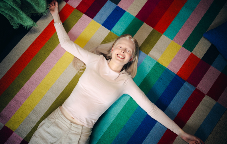 A blonde woman lies on her back on a colourful rug, arms spread wide and smiling to camera.