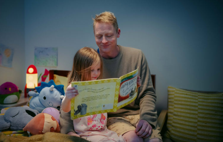 A father and daughter in their pyjamas are reading a bedtime story in the child’s bedroom.  