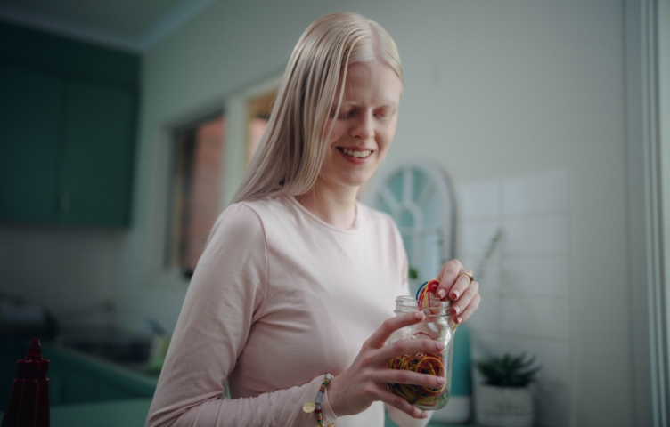 A blonde woman wearing a pink top stands in her kitchen, holding a jar filled with rubber bands.