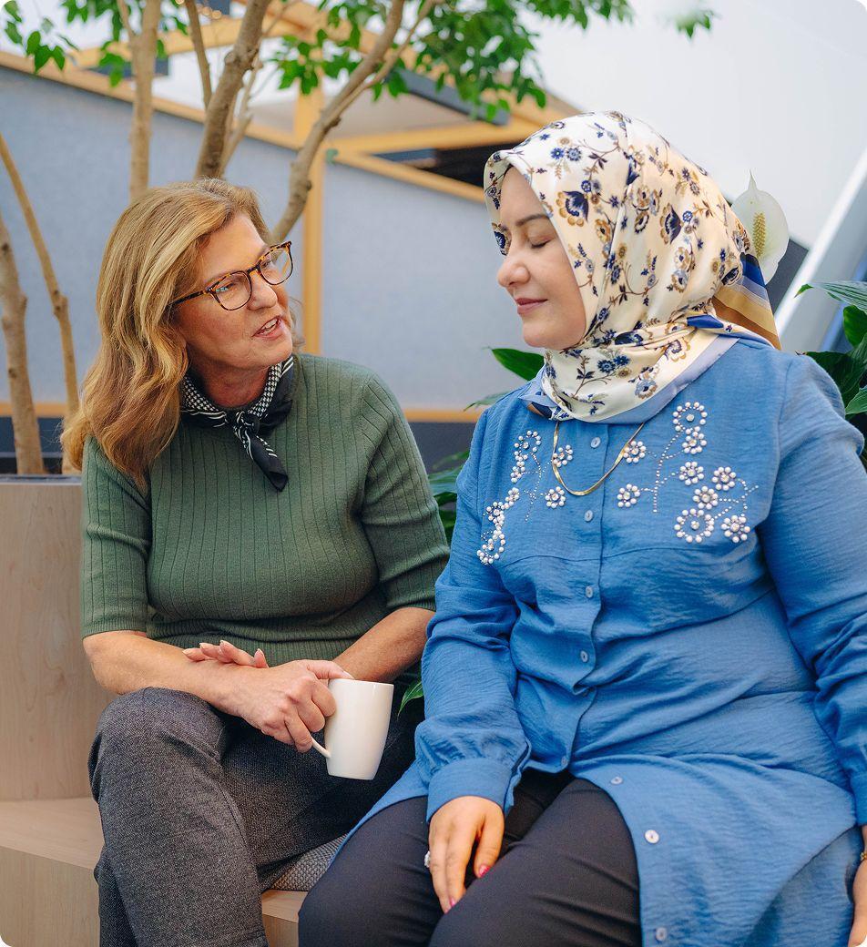 A female service provider drinks coffee with her client while sitting together on an indoor bench seat.