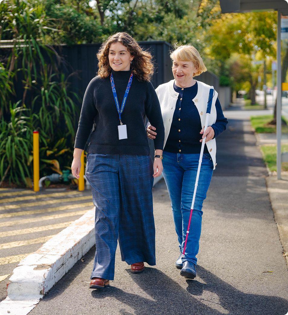 A female service provider walks with her female client outdoors, who is holding a white cane.