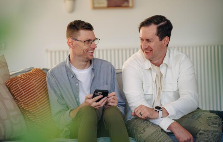 Two men sit on the couch smiling, reading something off a phone.